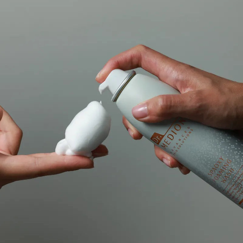 Person holding a spray bottle labeled 'Spaoxy Foaming Wash' over another person's hand against a gray background